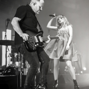 Black and white image of Iain playing the guitar and lauren dancing in the background