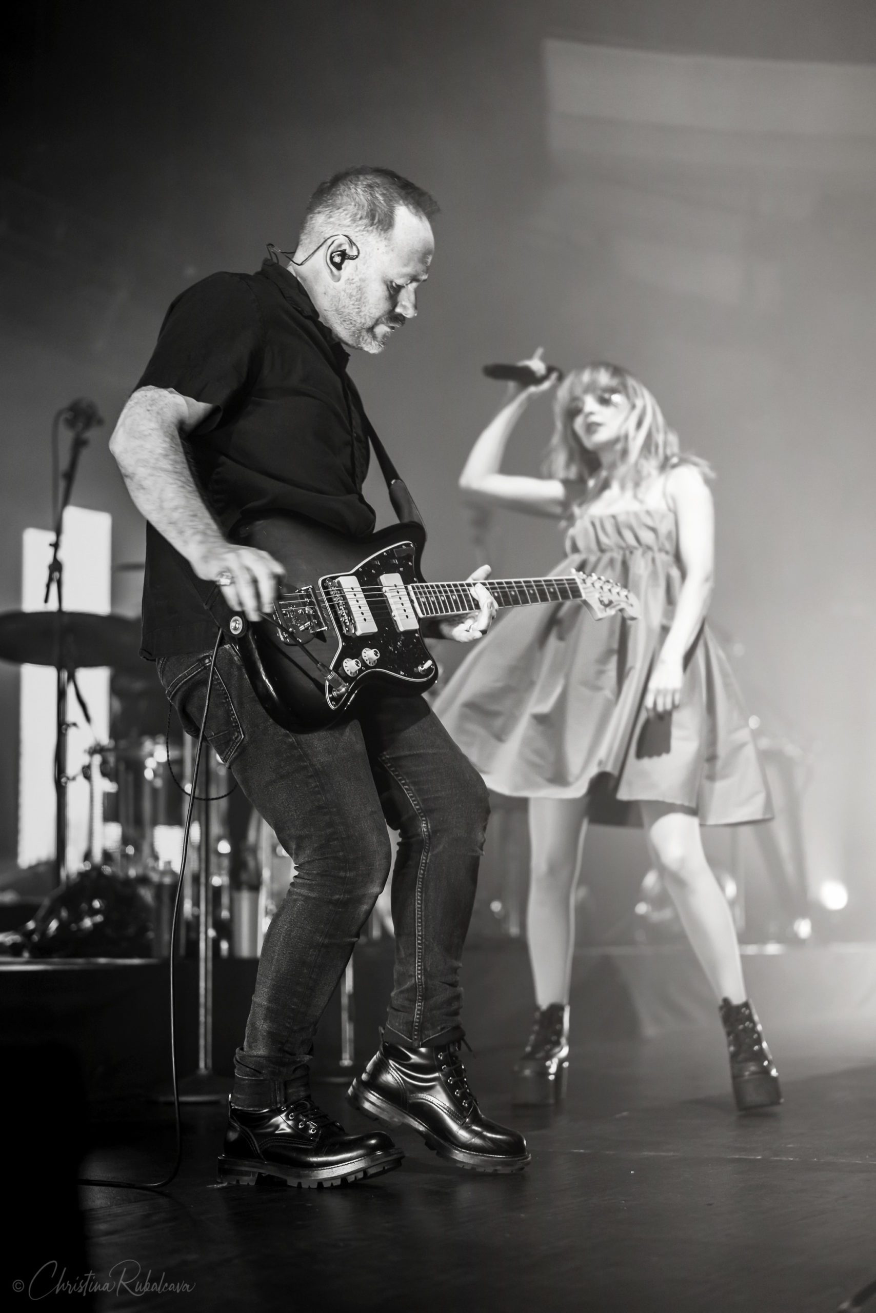 Black and white image of Iain playing the guitar and lauren dancing in the background