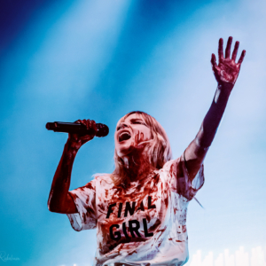 Lauren Mayberry singing into the microphone wearing a white T-shirt that says Final Girl on it and covered in fake blood.