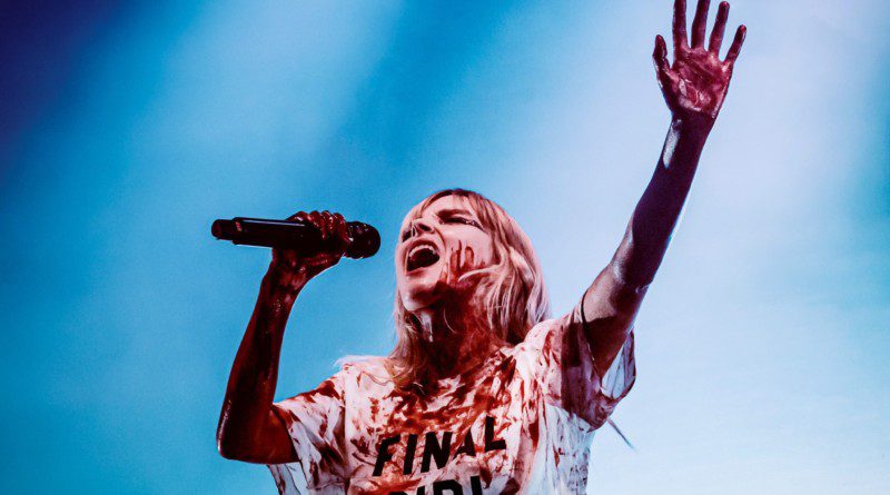 Lauren Mayberry singing with her hand raised, wearing a white shirt that says "Final Girl" covered in fake blood