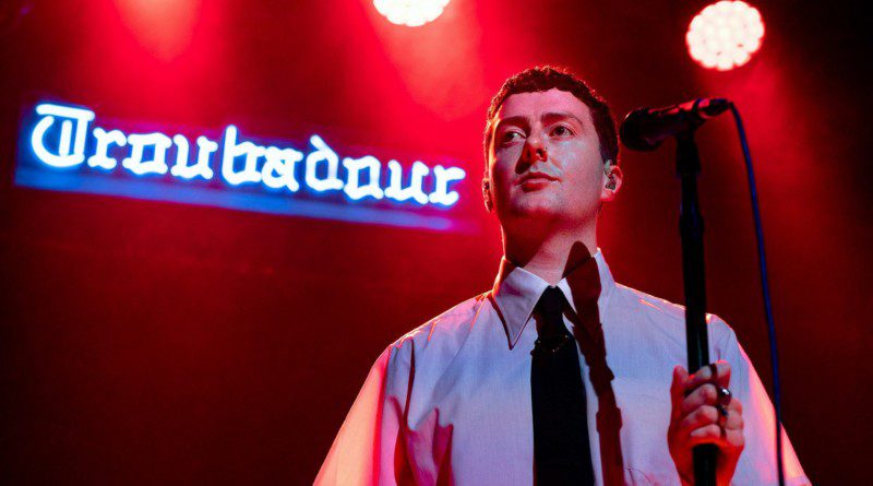 Joesef in a white short sleeve button up with a black tie and the neon sign in the background read "Troubadour"