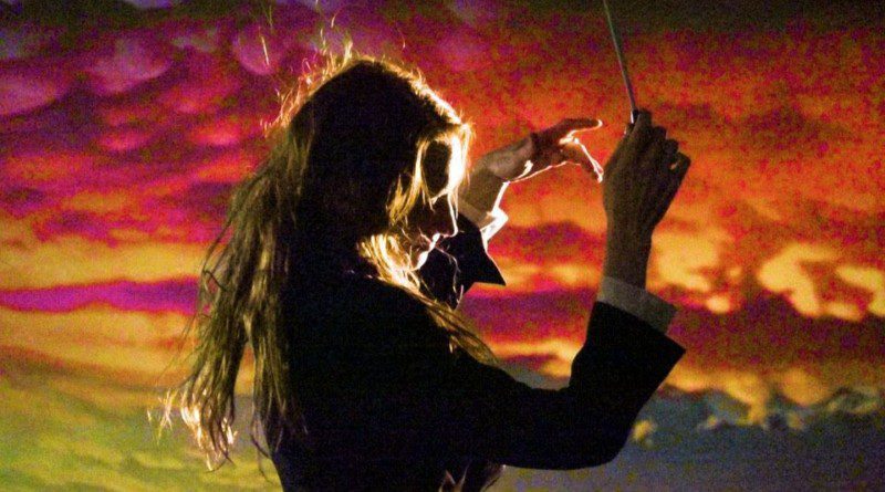 Melody English in front of a sunset looking background. You can see her side profile and She is wearing a black suit and white shirt with a conductor wand in hand.