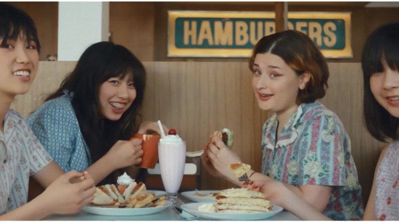 The Linda Lindas sitting at a table at a diner with burgers and milkshakes