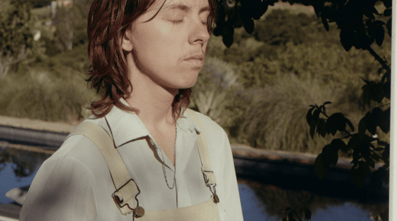 Meg Dufy with their eyes closed outdoors, trees and water in the background. Meg is wearing an off white button up shirt with tan colored overalls