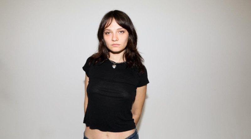 Clementine Creevy against a white backdrop with her hands behind her back. Clementine is wearing a black crop top tee and blue jeans.