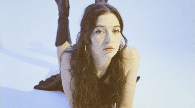 A pale blue backdrop and Alaska Reid laying down on her stomach and leaning on her arms and is looking at the camera. She is wearing a black dress and and black cowboy boots and her hair is dark brown and wavy which she is wearing in a half up pony tail