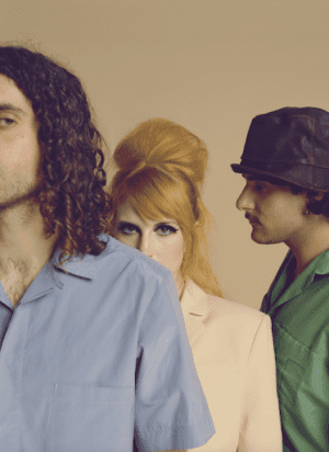 light yellowish background. Taylor york of paramore standing on the left with only half of his face visible. Hayley is standing in the middle peering over taylors shoulder and Zac is standing on the right of the image and his side profile is visible.