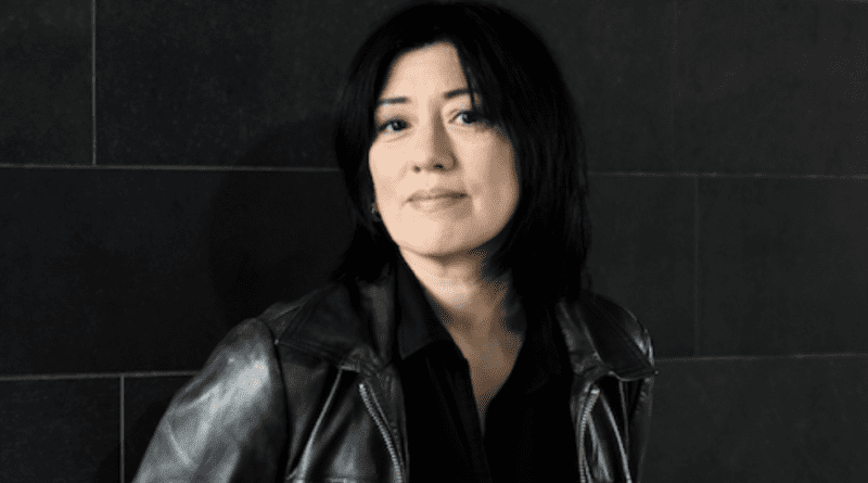 Miki Berenyi portrait. Miki is wearing a black leather moto style jacket, black top and is against a black background