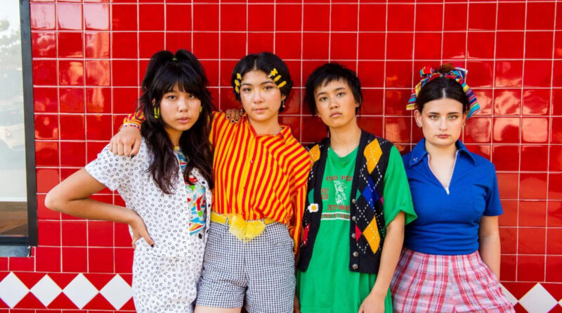 The band The Linda Lindas Standing in a row in front of a red tile wall